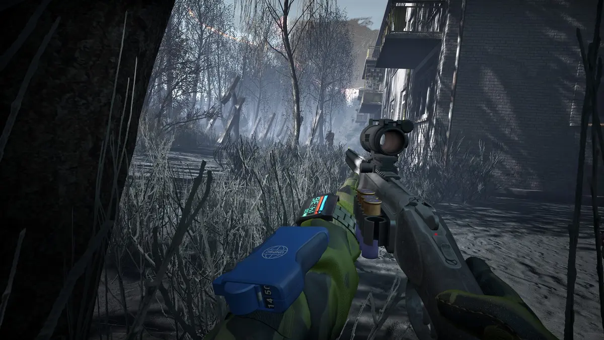 A first-person view in Into the Radius 2: a gloved hand holds a scoped pistol with a spare magazine over frosted reeds, with ruined tower blocks and a smokestack fading into grey mist.