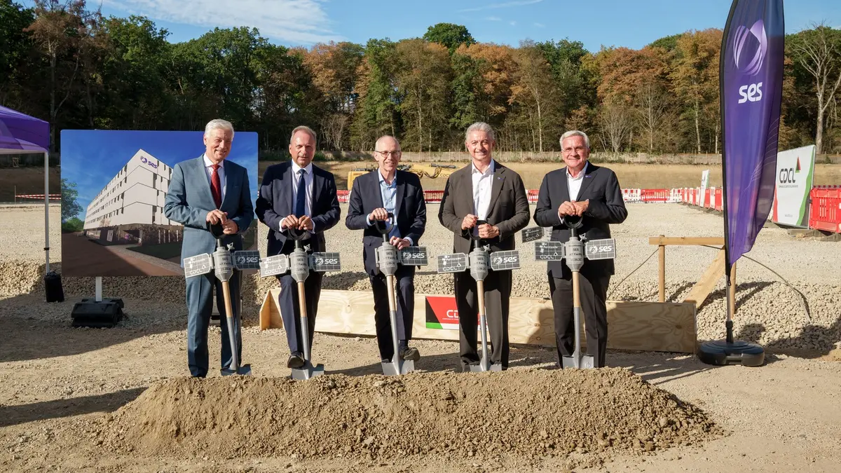 Five officials in suits holding ceremonial shovels in a mound of earth at a groundbreaking ceremony, with a large board showing a rendering of the planned building beside them, an SES branded flag and a partner banner on the right and a cleared construction site behind.