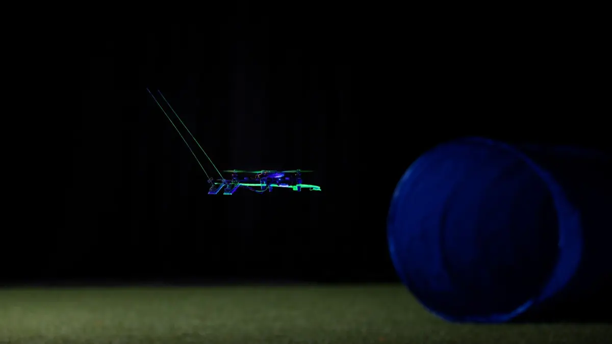 A small quadcopter carrying two long forward-pointing whisker rods hovers in near darkness above a dimly lit floor, with a large blue-lit pipe to the right of the frame