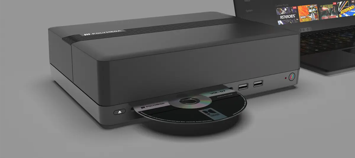 A Polymega Base Unit console with its disc tray open and a game disc fitted, a laptop showing the Polymega library screen behind it