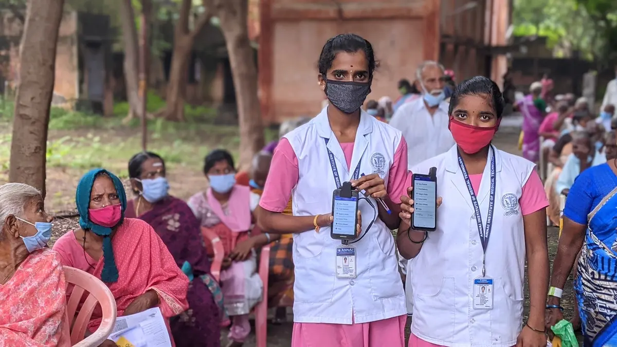 Two health workers from the All India Institute of Medical Sciences in masks and white coats holding smartphones showing a triage app, with patients waiting on plastic chairs behind them