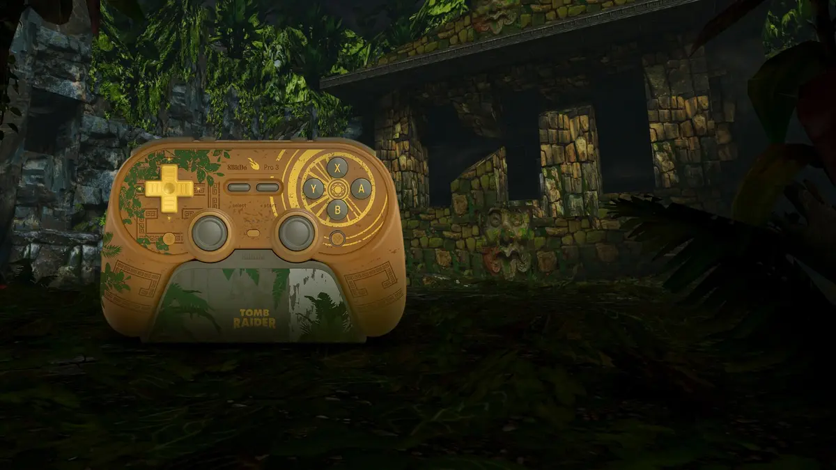 The sandstone-coloured 8BitDo Pro 3 Tomb Raider 30th Anniversary Edition gamepad, its gold D-pad and buttons picked out against creeping vine patterns, resting in overgrown jungle ruins