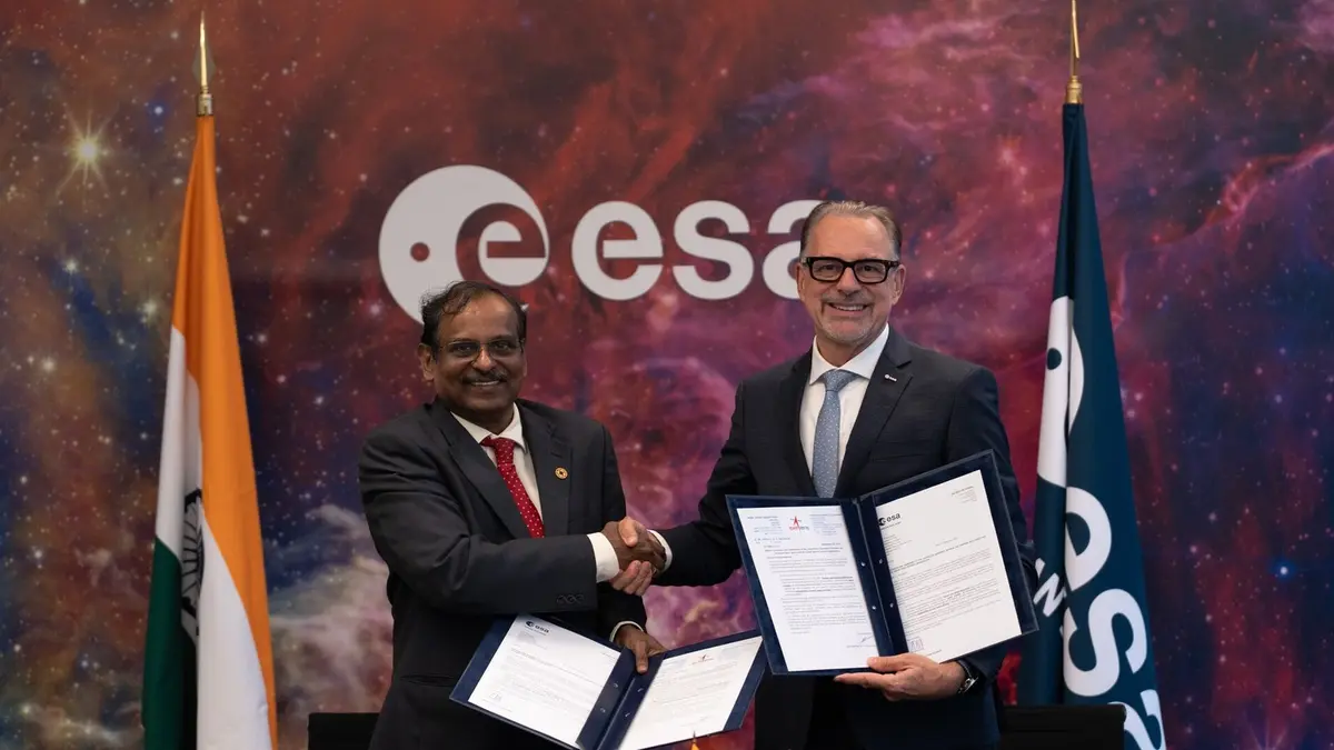 Two officials shake hands while holding signed document folders, with the ESA logo behind them, the Indian flag on the left and the ESA flag on the right, against a backdrop of stars and nebula clouds.