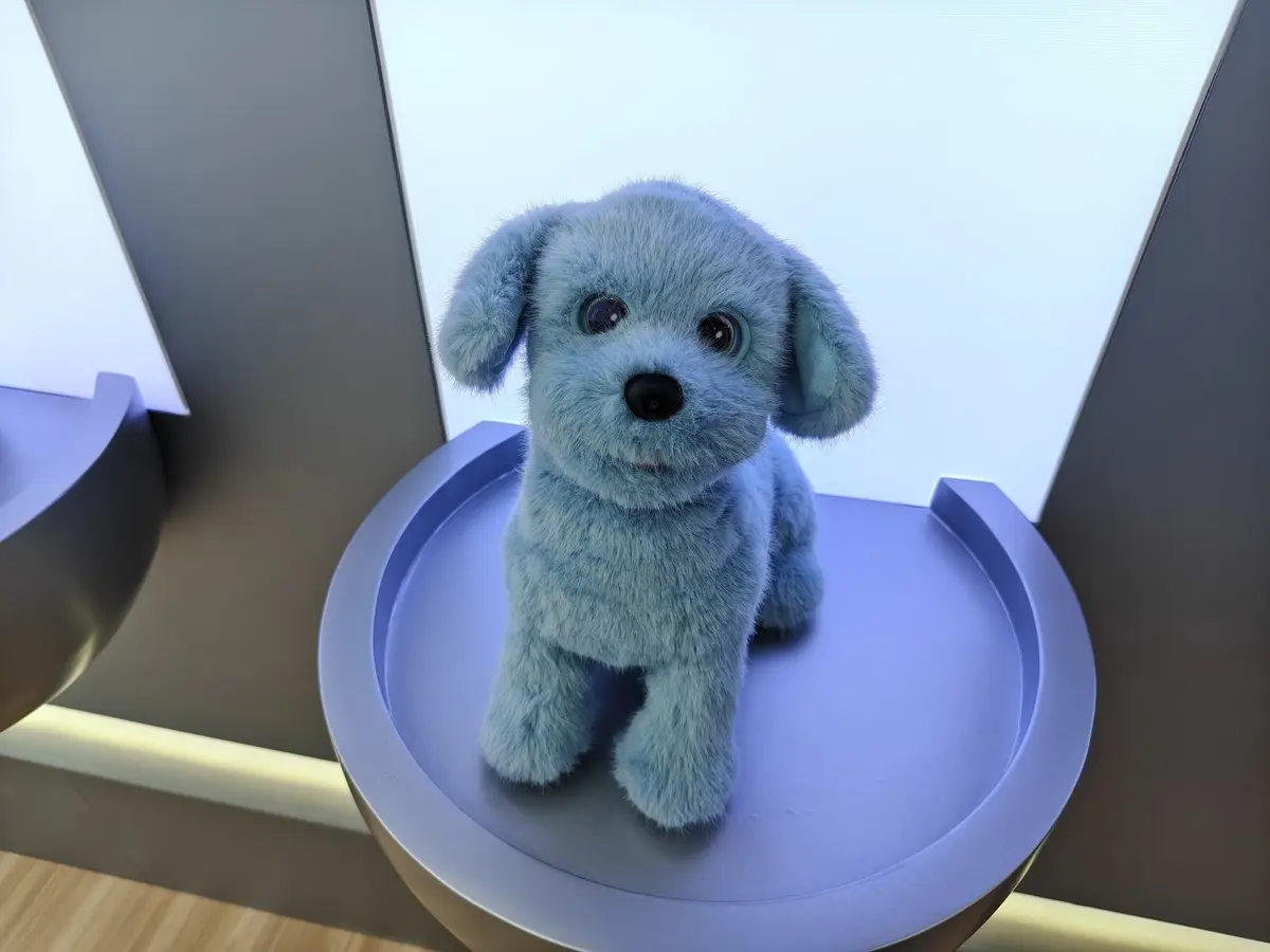 Lilmilo AI companion robot photographed at IFA 2026: a small blue plush-like robot with floppy ears, a black nose and large glossy eyes on a lavender display platform.