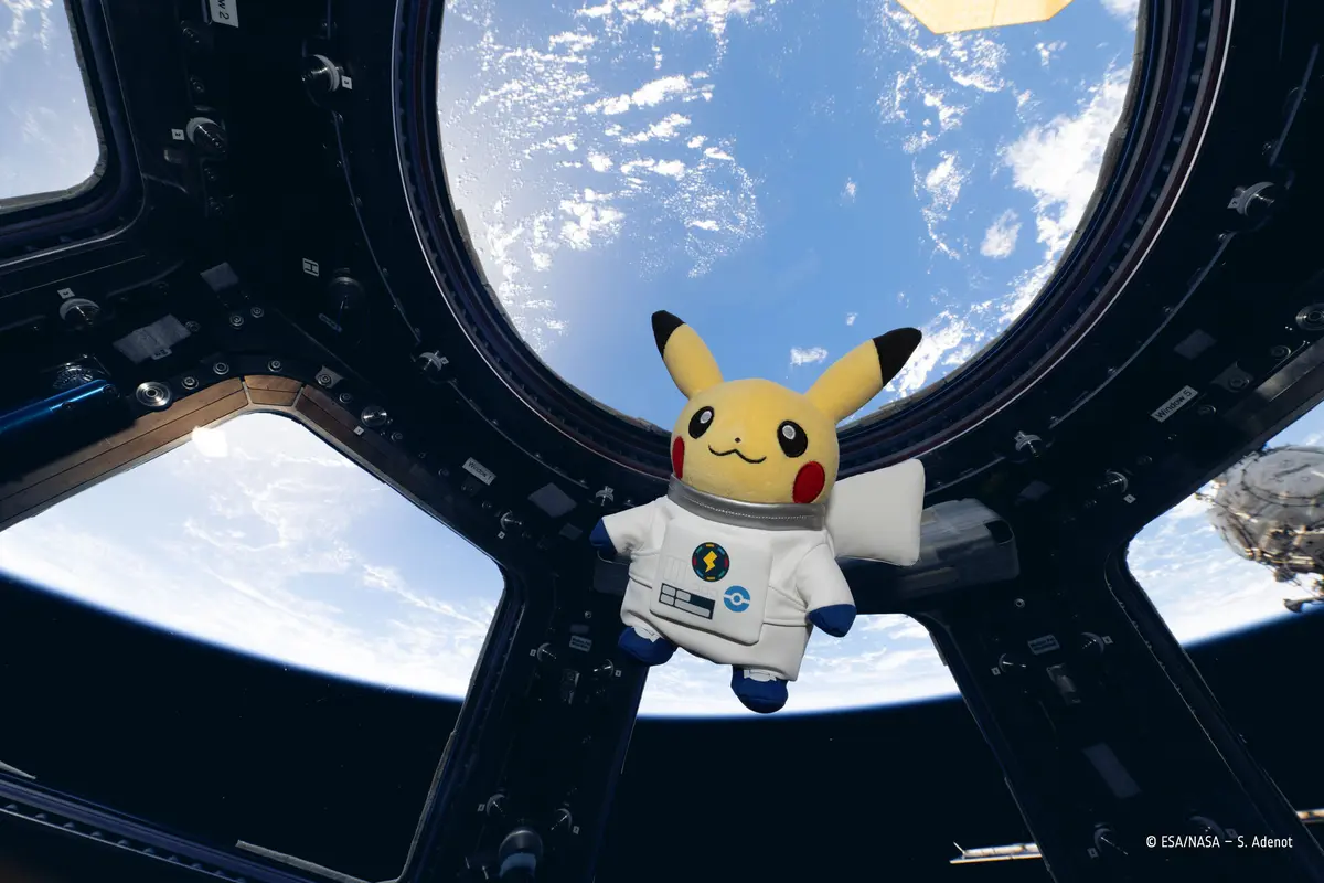 A Pikachu plush wearing a white astronaut suit with an ESA mission patch floating inside the cupola of the International Space Station, with Earth's blue oceans and white clouds visible through the surrounding windows