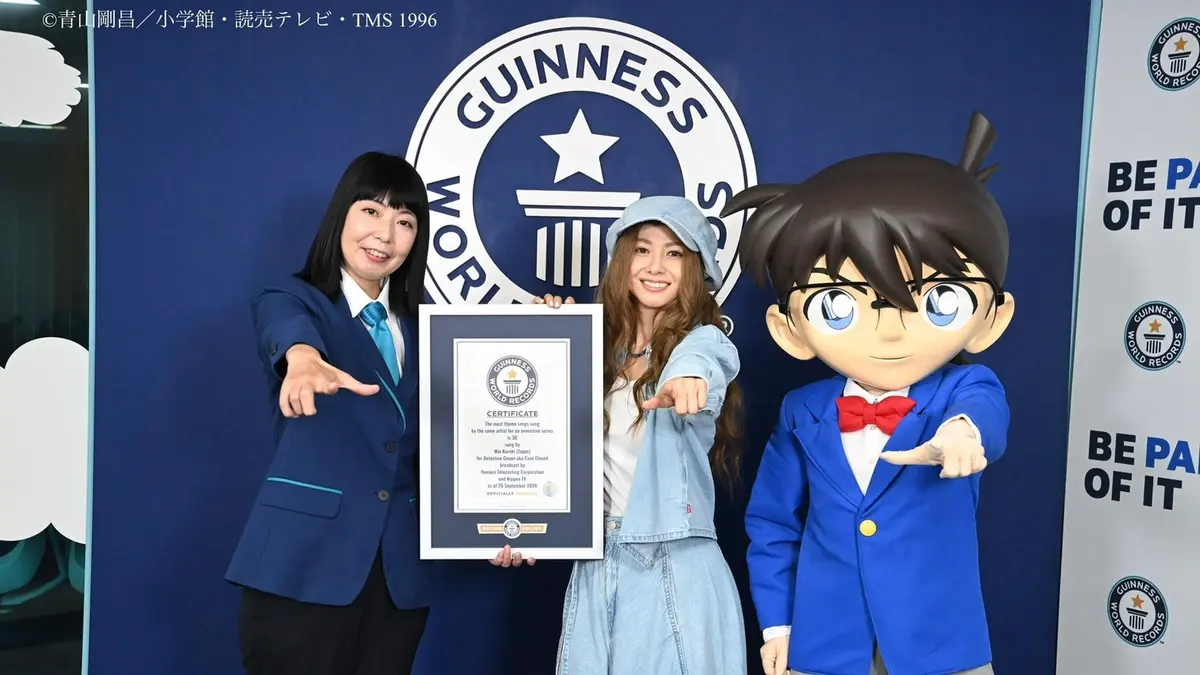Guinness World Records certificate presentation: Mai Kuraki in a denim jacket, jeans and cap holds the framed certificate for 30 Detective Conan theme songs, flanked by an adjudicator in a navy suit and a Conan Edogawa mascot in a blue blazer and red bow tie, all three pointing at the certificate against a deep blue Guinness World Records backdrop