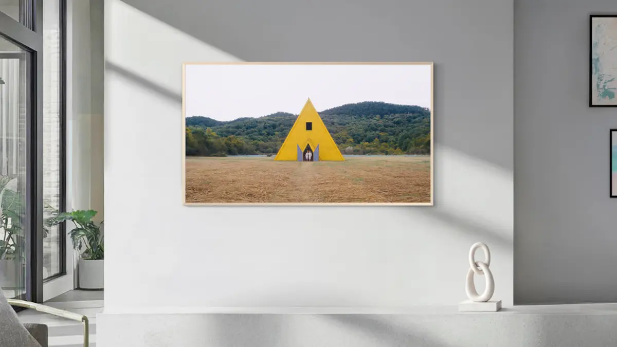 A Samsung The Frame television on a pale wall showing a yellow pyramid structure in a green field, with a plant and a small sculpture in the room around it