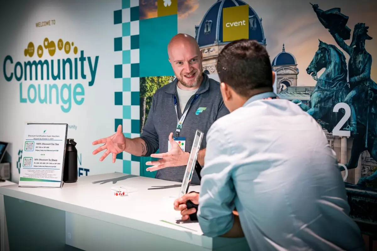 Two people in conversation across a table in the Community Lounge of a Microsoft data conference, with banners and exhibition stands behind them