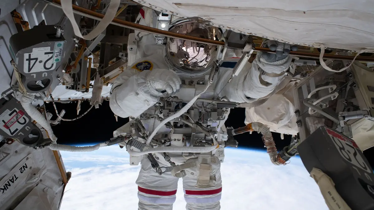 An astronaut in a white spacesuit with red stripes on the legs works on the outside of a space station, one gloved hand gripping a rail as the curved horizon of Earth and the cloud tops below run across the lower half of the frame, with the black of space behind.