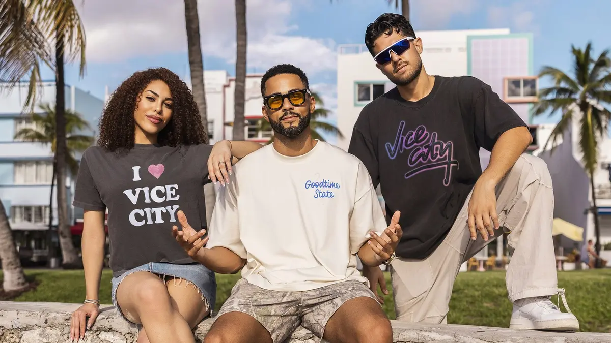 Three people in Miami HEAT Vice City collection T-shirts and shorts sit in a sunlit Miami park, with pastel art-deco buildings and palm trees behind them