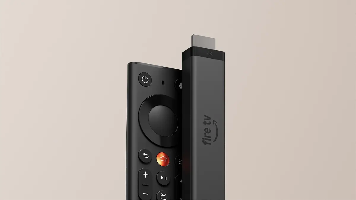The Fire TV Stick 4K standing upright beside Amazon's new Fire TV Remote, showing the remote's circular direction pad, metal home button and volume keys against a plain beige studio background