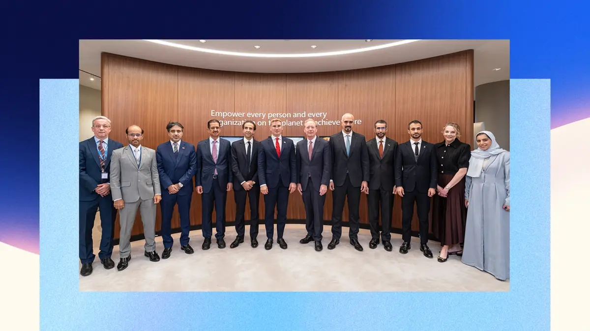 A group photograph of Microsoft executives and Gulf officials standing in a row in front of a curved wooden wall carrying Microsoft's mission statement, shown inside a blue gradient frame.