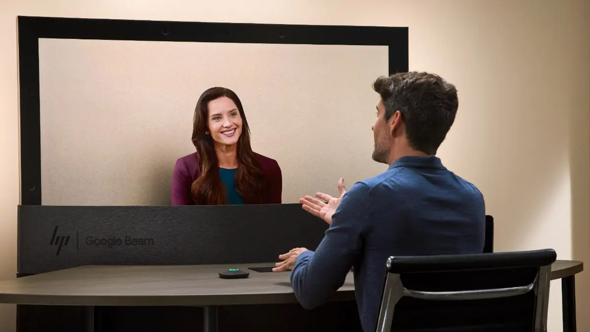 An HP Dimension with Google Beam video booth in an office, with the HP and Google Beam wordmarks on the console, a woman visible on the booth screen and a man seated at a table talking to her.