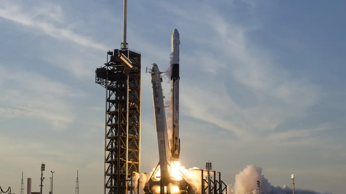 A SpaceX Falcon 9 rocket carrying a Crew Dragon capsule climbs away from Launch Complex 39A at NASA's Kennedy Space Center, the launch tower standing beside it and exhaust spreading across the pad beneath a pale blue sky.