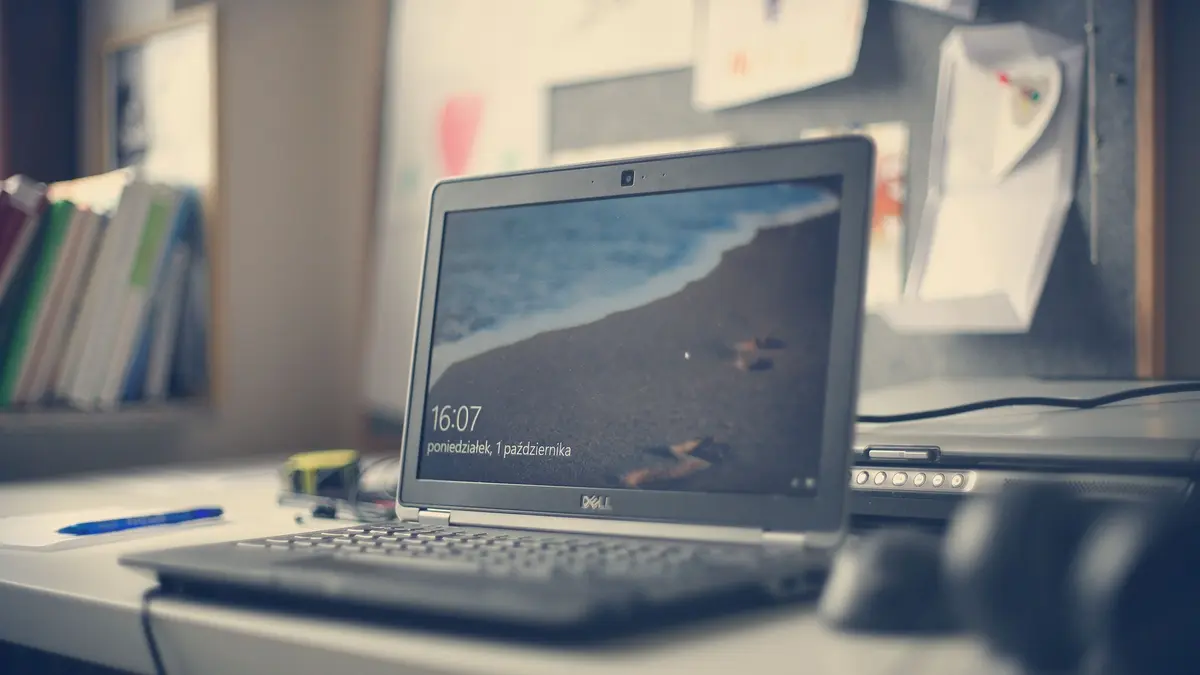 A silver laptop open on a desk showing its lock screen, with notebooks, a pen and papers behind it.