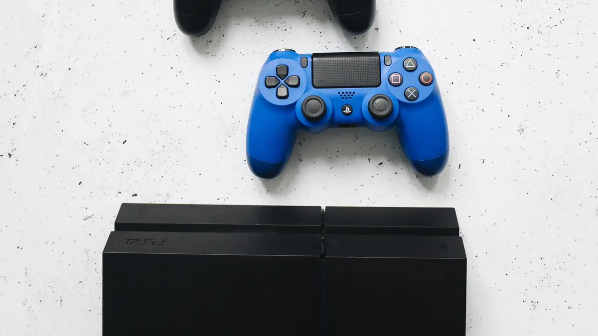 A blue DualShock 4 controller lying above two PlayStation 4 consoles on a white speckled surface.