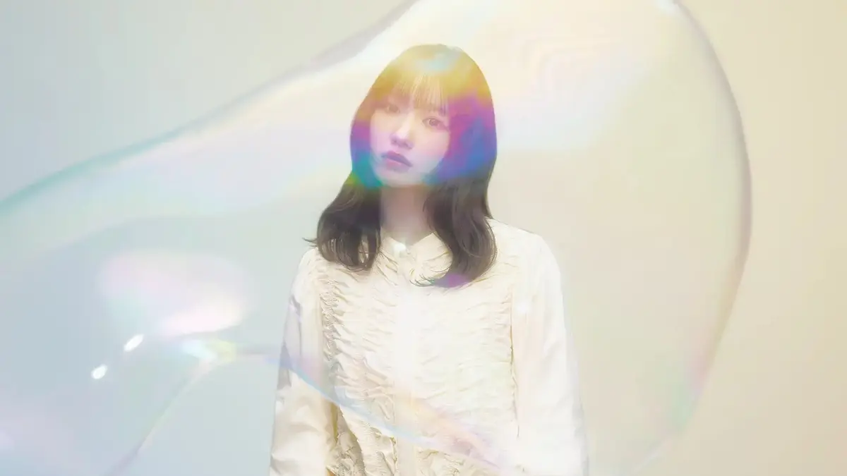 Yuika stands against a pale studio wall with a large iridescent soap bubble drifting across her face, in the portrait published with the announcement that she sings the opening theme of the Promise Me the Spotlight anime.