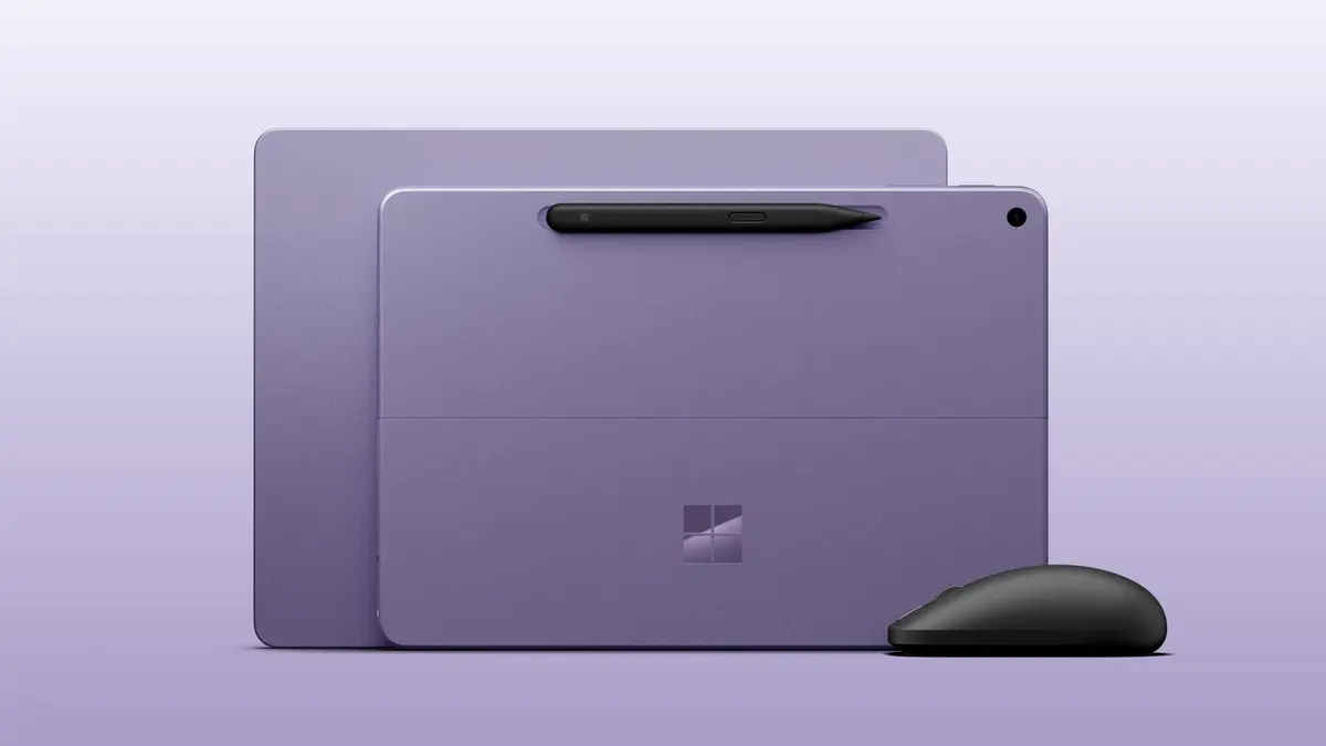Microsoft’s press image of two purple Surface devices standing side by side, with a black Slim Pen clipped to the front tablet’s keyboard cover and a black Surface Mouse in front of it on a lavender background.