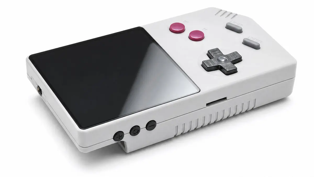 Angled product shot of the FPGB Mini handheld: a small grey console with a black screen, a black cross-shaped D-pad and two magenta face buttons, standing on a white surface.