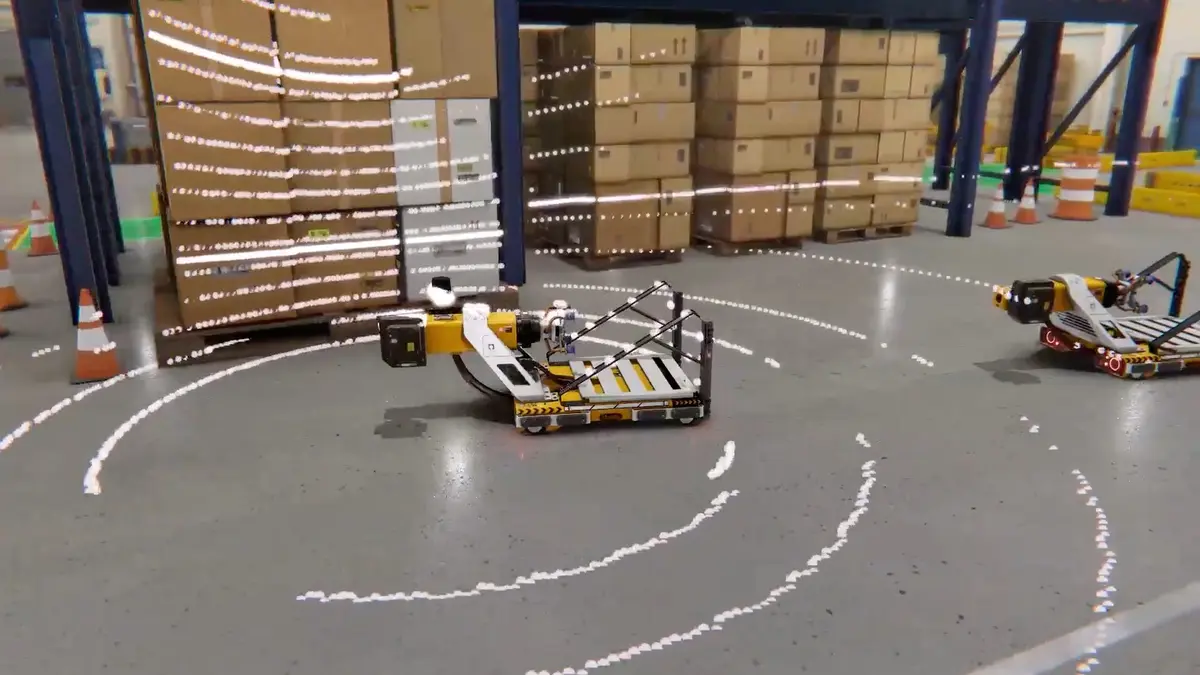 Two small wheeled robots on a grey warehouse floor, a white dotted sensor-scan pattern sweeping across the concrete around them, with stacked cardboard cartons and traffic cones behind.