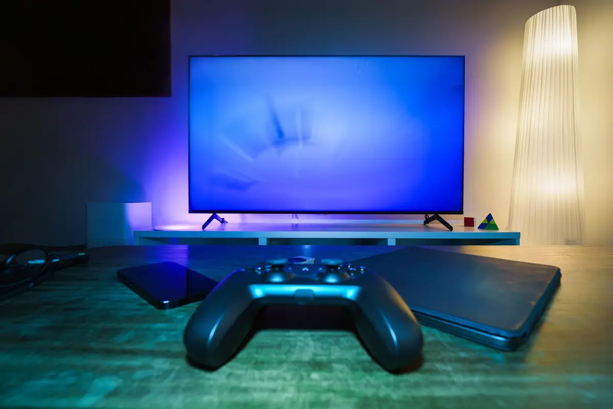 A television screen glowing blue in a living room, with a wireless games controller, a phone and a laptop on the table in front of it.