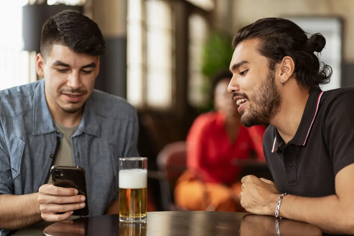 Two men sit at a bar table with a pint of beer as one checks his smartphone, illustrating digital proof of age for alcohol sales.