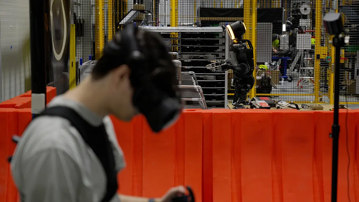 An Atlas humanoid robot stands inside a fenced robot cell in a car plant, its black bodywork topped by a glowing amber head, while a worker wearing a head-mounted display operates equipment in the foreground behind an orange safety barrier.