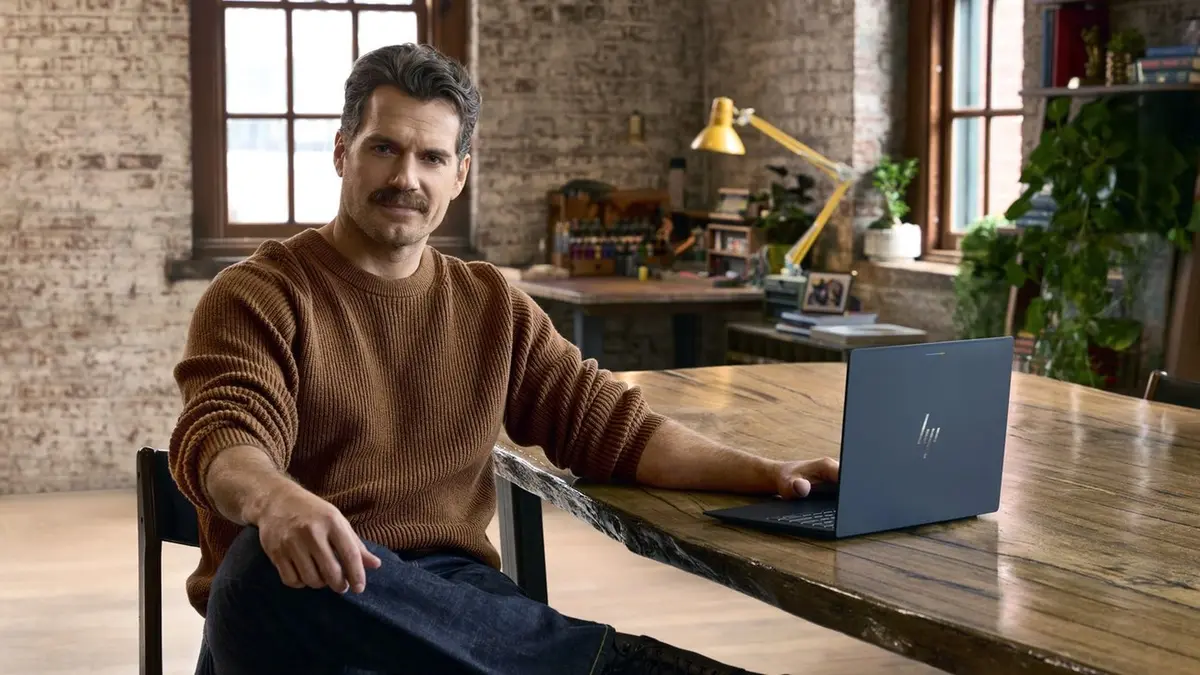 Henry Cavill in a brown ribbed jumper, sitting at a wooden table with a dark grey Googlebook laptop beside him, in a brick-walled studio with plants and a yellow desk lamp behind