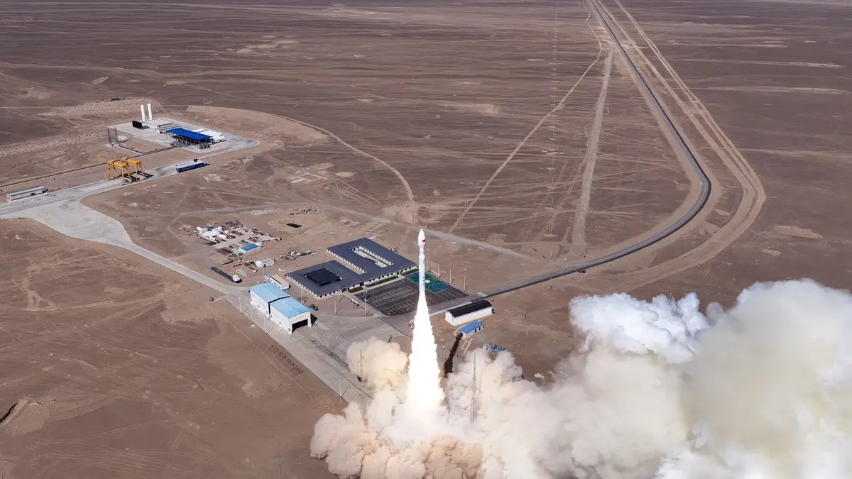 An aerial view of a Kinetica 1 rocket climbing away from a desert launch complex, its exhaust plume spreading across the ground beside low service buildings, a concrete apron and a rail line