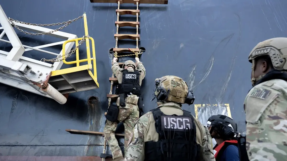 Coast Guard boarding team members in camouflage and helmets climbing a rope ladder up the dark hull of a tanker at sea while colleagues in body armour wait on a small boat below