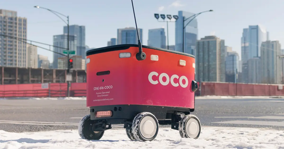 A Coco Robotics delivery robot on a wintry city street, with high-rise buildings and traffic lights behind it