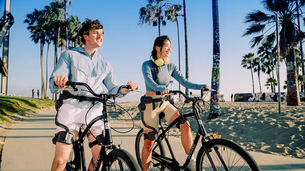 Two people riding bicycles while wearing Ascentiz wearable exoskeleton modules around their hips and knees.