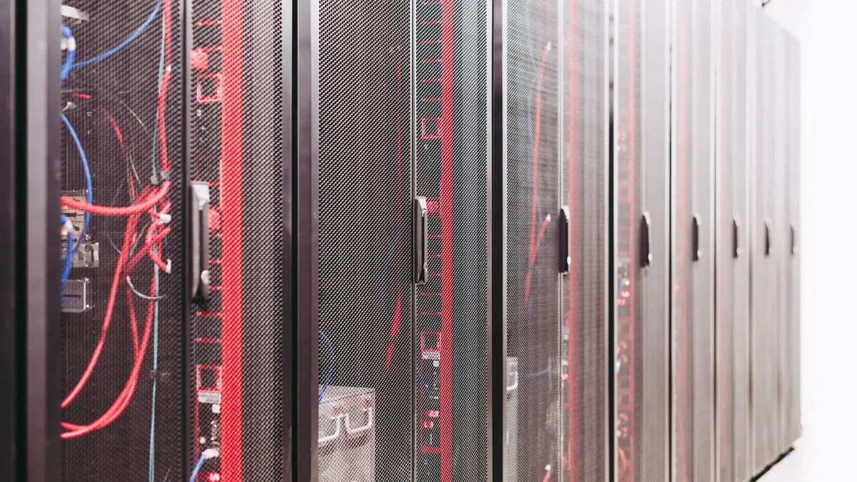 A row of mesh-fronted server racks in a data centre aisle, with red and black cables running down the nearest cabinet and pale flooring visible at the far end.