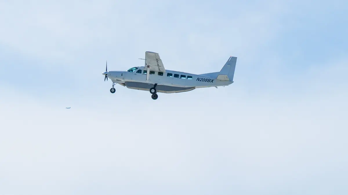 A white twin-turboprop NASA research aircraft with its landing gear extended on final approach, the registration N208BX visible on its rear fuselage, against a pale blue sky.