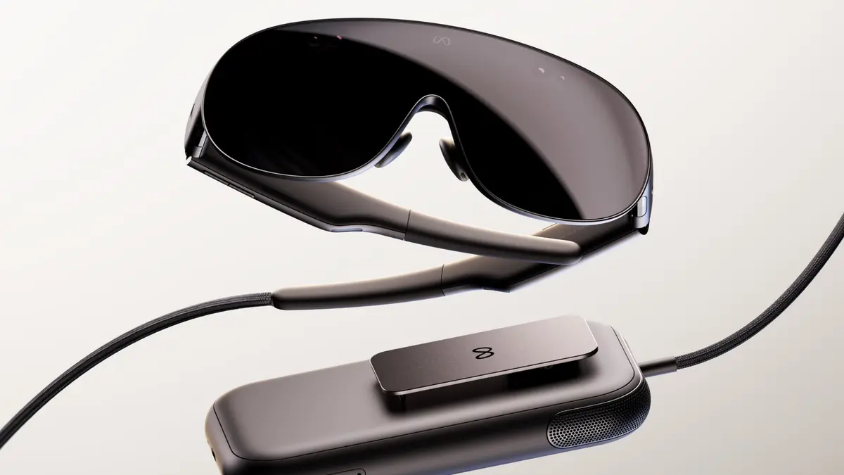 Meta’s VR Glasses shown with the clip-on compute puck: a dark wraparound visor with the Meta logo on the lens, joined by a braided optical tether to a rounded grey puck, also carrying the Meta logo, on a pale background.