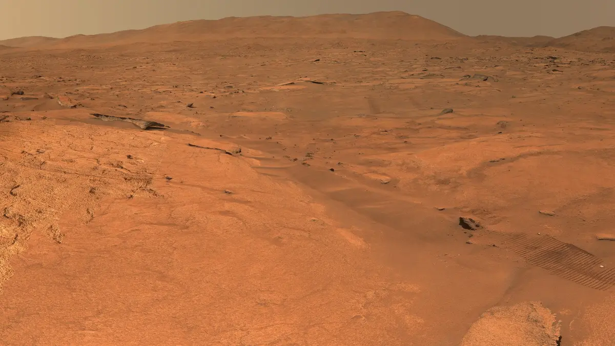 A wide view across rust-coloured Martian ground from Perseverance: a rocky outcrop on the left, pale bedrock ledges in the middle distance, wheel tracks crossing the slope on the right and a dust-hazed tan sky above the hills on the crater rim.