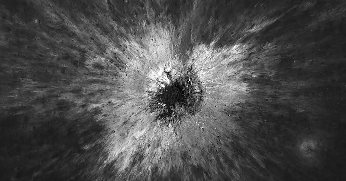 A fresh impact crater on the Moon seen from orbit: a dark, roughly circular pit ringed by bright streaks of ejected material radiating outwards across the grey surface.