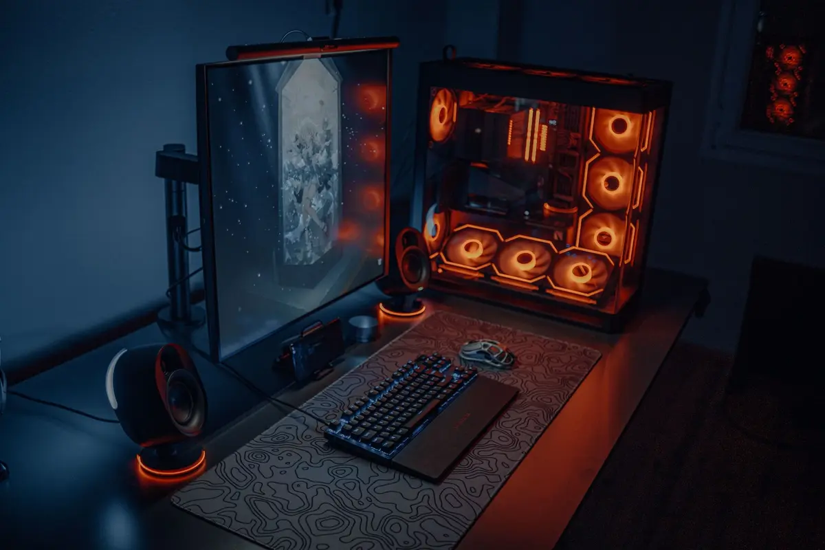 A gaming desk at night with a widescreen monitor, mechanical keyboard and a glass case PC lit by purple RGB fans