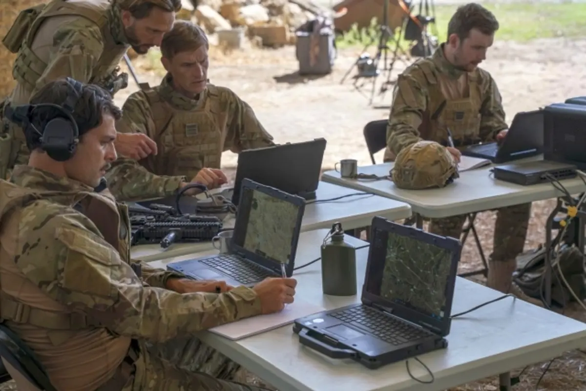 Soldiers in camouflage uniforms working at laptops on folding tables in a field command post, with helmets and radio equipment laid out beside them
