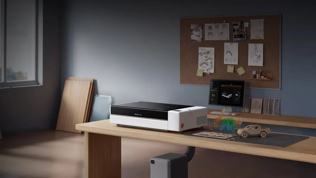A white and black Bambu Lab R1 laser cutter with its lid closed on a wooden workbench in a home workshop, beside 3D-printed models and a sheet of cardboard being cut, with timber boards leaning against the wall behind and a monitor showing the Bambu Suite layout