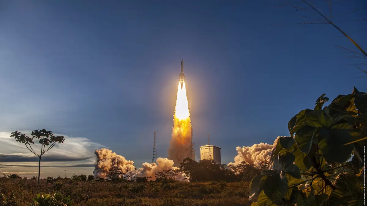 An Ariane 6 rocket lifting off from the Guiana Space Centre at dusk, its bright exhaust plume lighting thick clouds of smoke between two launch towers, with dark foliage framing the foreground.