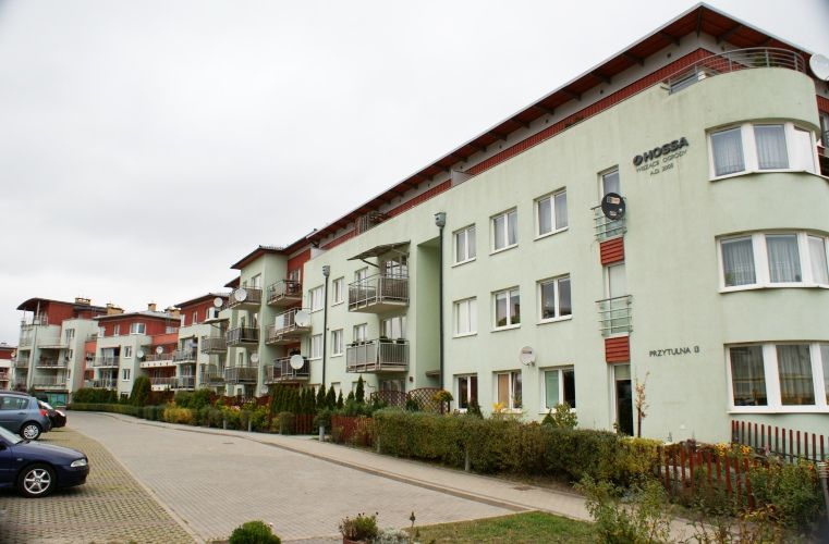 Szereg nowoczesnych budynków mieszkalnych z jasnozielonymi fasadami, czerwonymi dachami, balkonami, wybrukowaną ulicą z zaparkowanymi samochodami i zielenią pod zachmurzonym niebem.