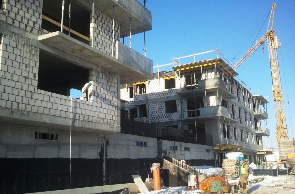 Plac budowy z wielopiętrowymi budynkami w trakcie wznoszenia, żółtym dźwigiem wieżowym i pracownikiem na zaśnieżonym gruncie.