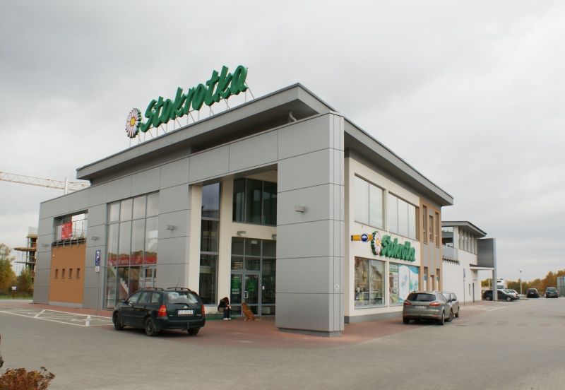 Nowoczesny budynek supermarketu „Stokrotka” z dużym szyldem, zaparkowanymi samochodami i zachmurzonym niebem.