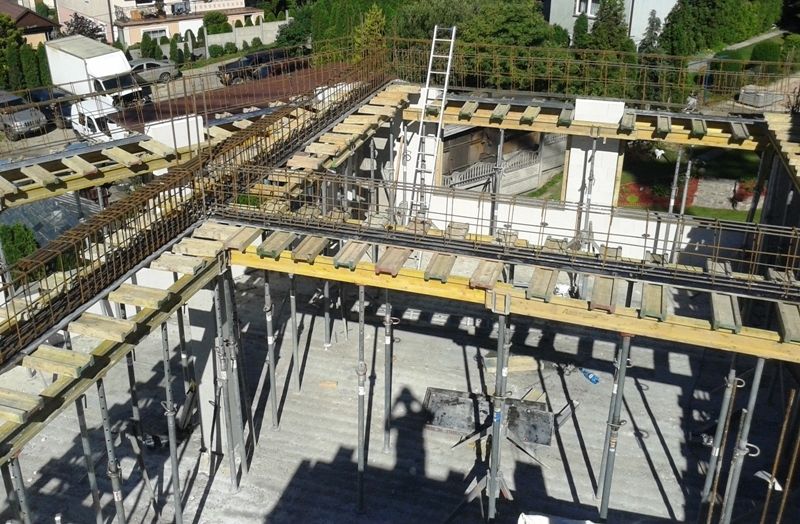 Widok z góry na budynek w trakcie budowy, ukazujący pręty zbrojeniowe, szalunki i metalowe podpory nowej podłogi.