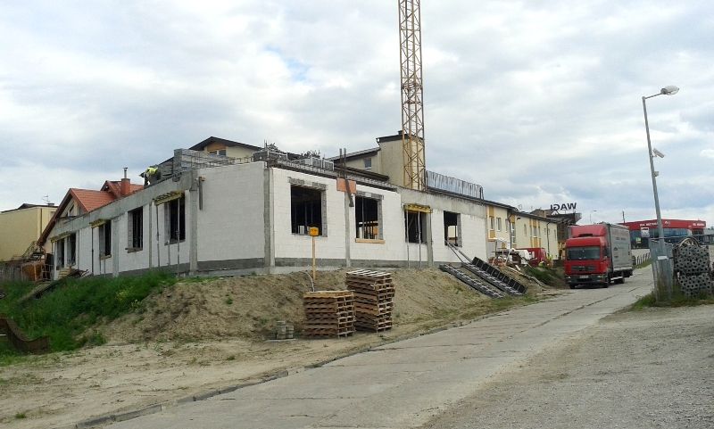 Budynek w budowie z odsłoniętymi betonowymi ścianami, kopcem ziemi, drewnianymi paletami, dźwigiem w tle i czerwoną ciężarówką na asfaltowej drodze.