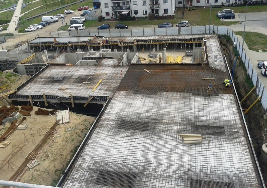 Plac budowy z odsłoniętymi betonowymi ścianami fundamentowymi i siatką zbrojeniową ułożoną na dużej płycie, z robotnikami.