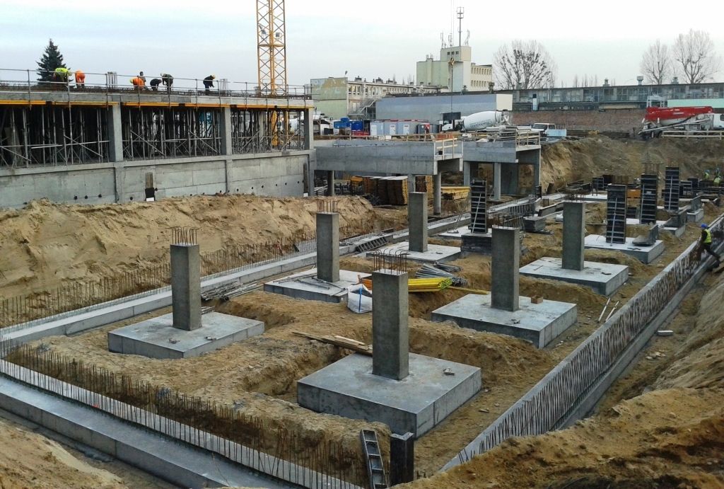 Plac budowy z wieloma betonowymi fundamentami i filarami wystającymi z wykopanej ziemi, w tle trwające prace budowlane.