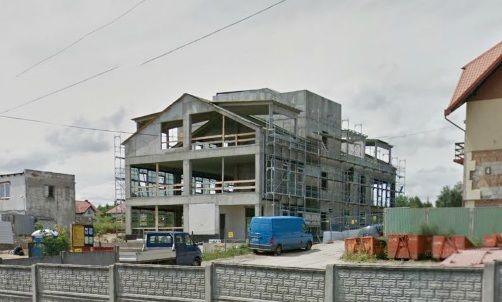 Duży betonowy budynek w budowie. Na pierwszym planie znajduje się rusztowanie, niebieski samochód dostawczy i materiały budowlane.