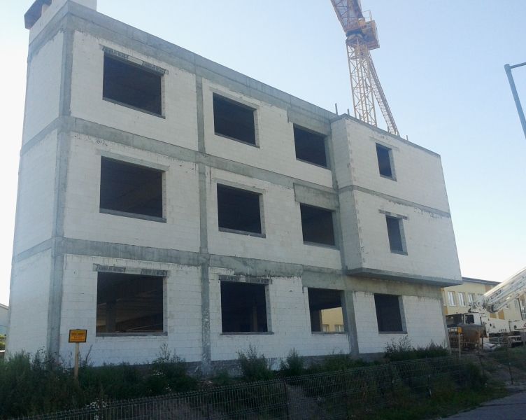 Trzypiętrowy budynek w budowie z białymi, bloczkowymi ścianami i odsłoniętymi betonowymi ramami. W tle dźwig budowlany.